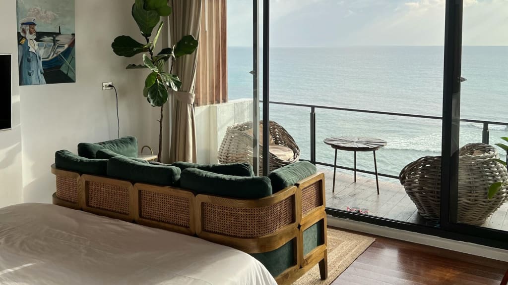 A sunlit room with a bed, green sofa, potted plant, and balcony overlooking the ocean. Wicker chairs and a small table are on the balcony.